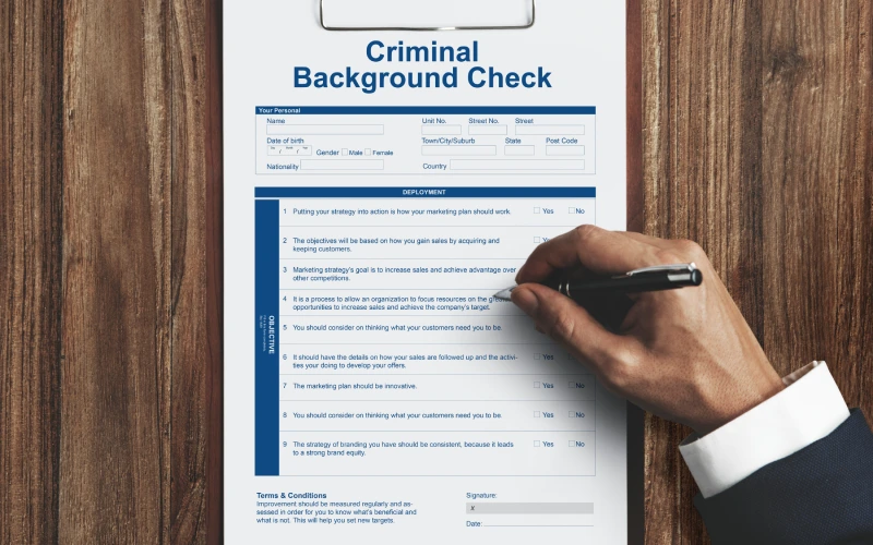 Person filling out a criminal background check form on a clipboard placed on a wooden desk